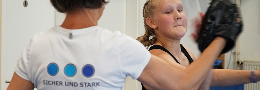 Ein Mädchen boxt gegen einen Schutzhandschuh, gehalten von einer Trainerin. Diese trägt ein T-Shirt mit der Aufschrift "Sichr & Stark"
