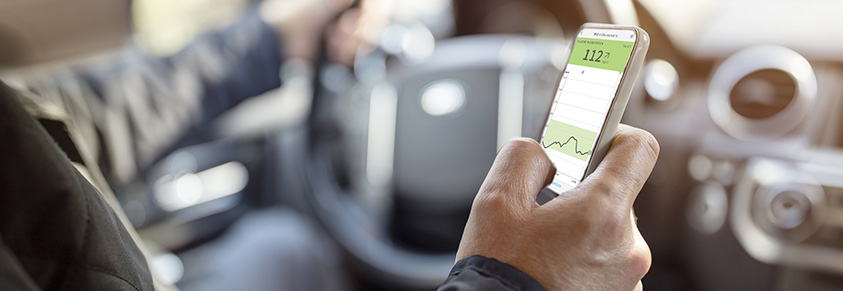 Ein Mann am Steuer schaut auf das Displays seines Handys um den Glucosewert auszulesen.