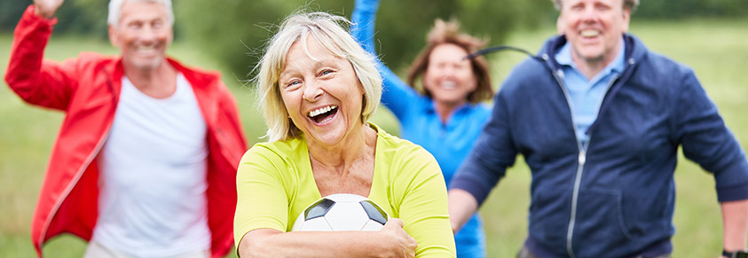 Eine ältere Dame hält einen Fußball in der Hand. Im Hintergrund freut sich eine Gruppe älterer Männer.