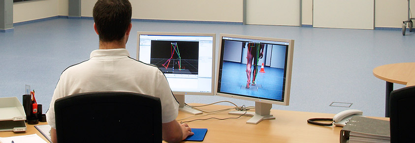 Ein Mann ist in einem Labor zu sehen von hinten am Schreibtisch, vor ihm zwei Monitore, die 3D-Modelle eines Gangbildes zeigen