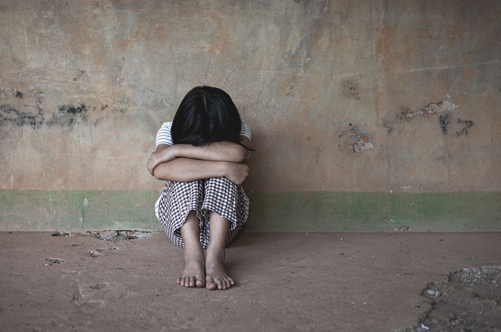 einsames Kind einsames, armes, obdachloses, verzweifeltes, traumatisiertes Kind, sitzt barfuß vor einer kargen, schmutzigen Wand, den Kopf auf die Knie gestützt, mit beiden Armen fest umschlungen