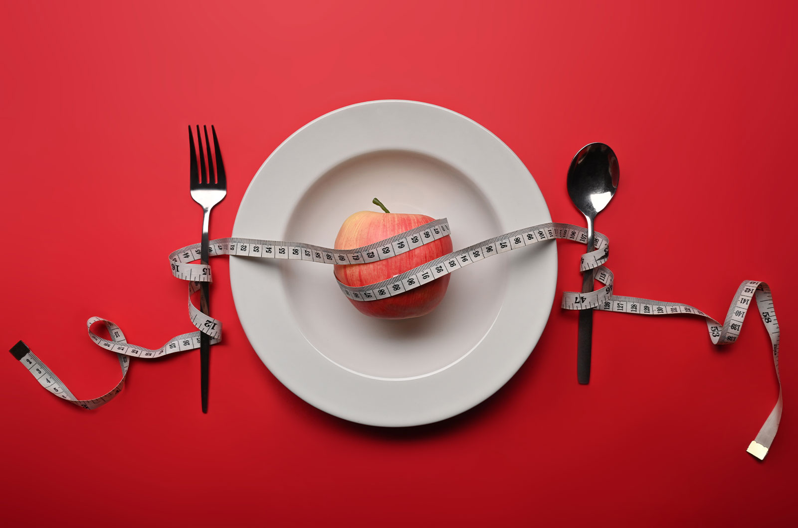 Apfel, Maßband auf einem Teller, Gabel und Löffel liegen daneben, Gewichtsabnahme mit einem gesunden Ernährungskonzept und Bewegung