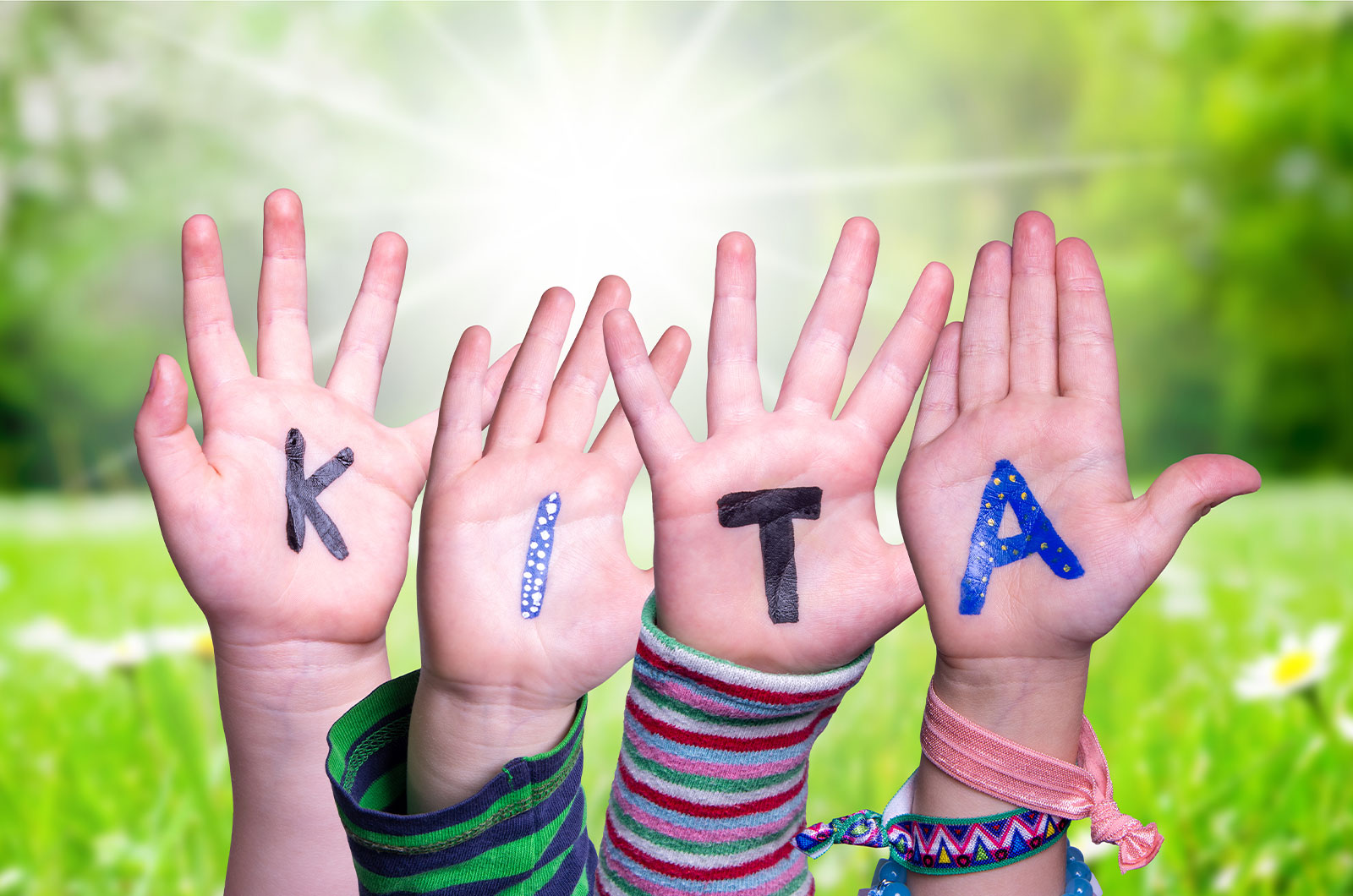 auf 4 kleinen Kinderhänden steht in farbigen Großbuchstaben das Wort KITA, als Hintergrund wurde eine sonnige grüne Graswiese gewählt