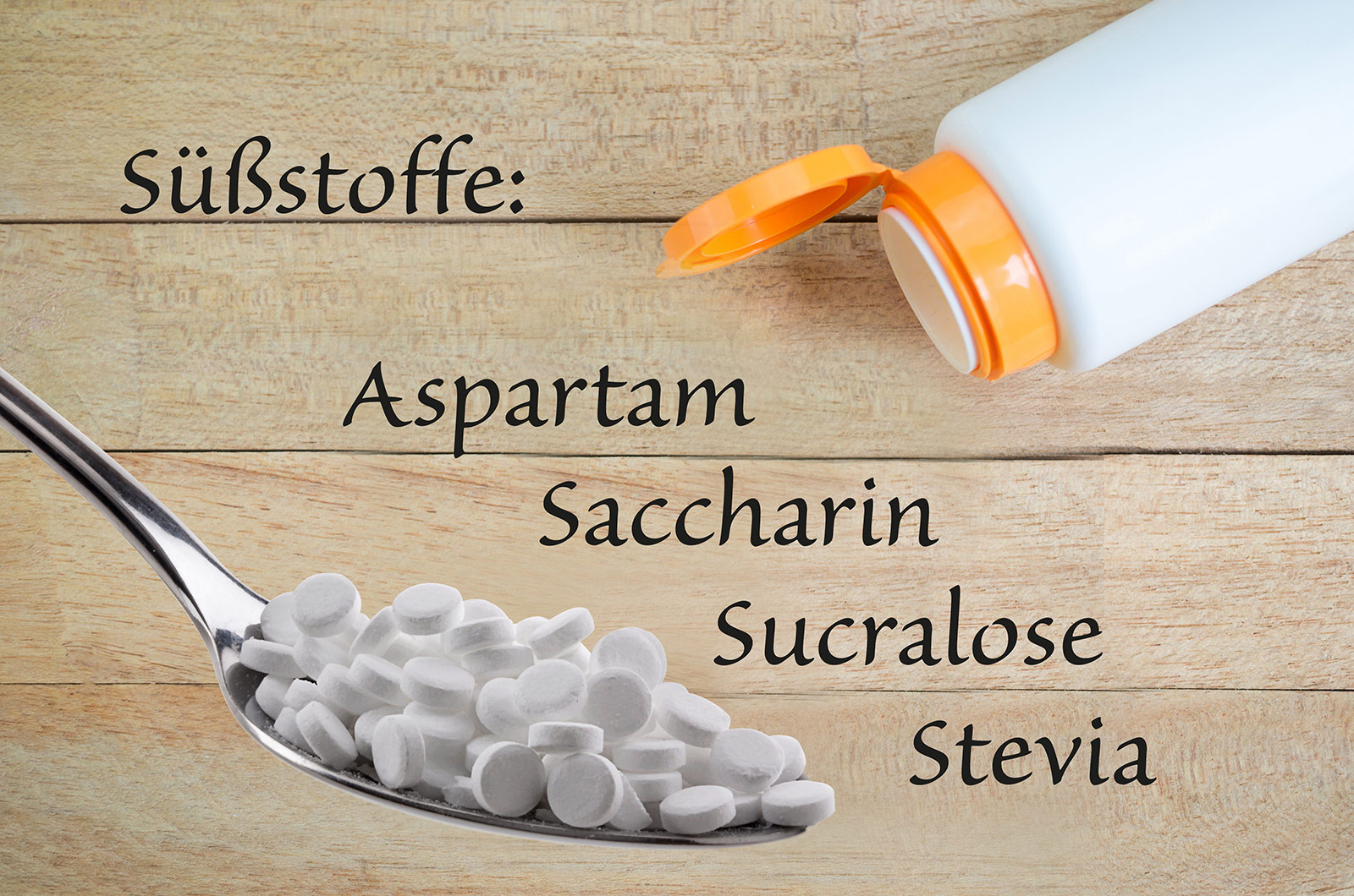 Süßstoff-Pillen auf einem Esslöffel, Pillendose auf  Holzhintergrund mit Beschriftung Süßstoffe: Aspartam, Saccharin, Sucralose und Stevia