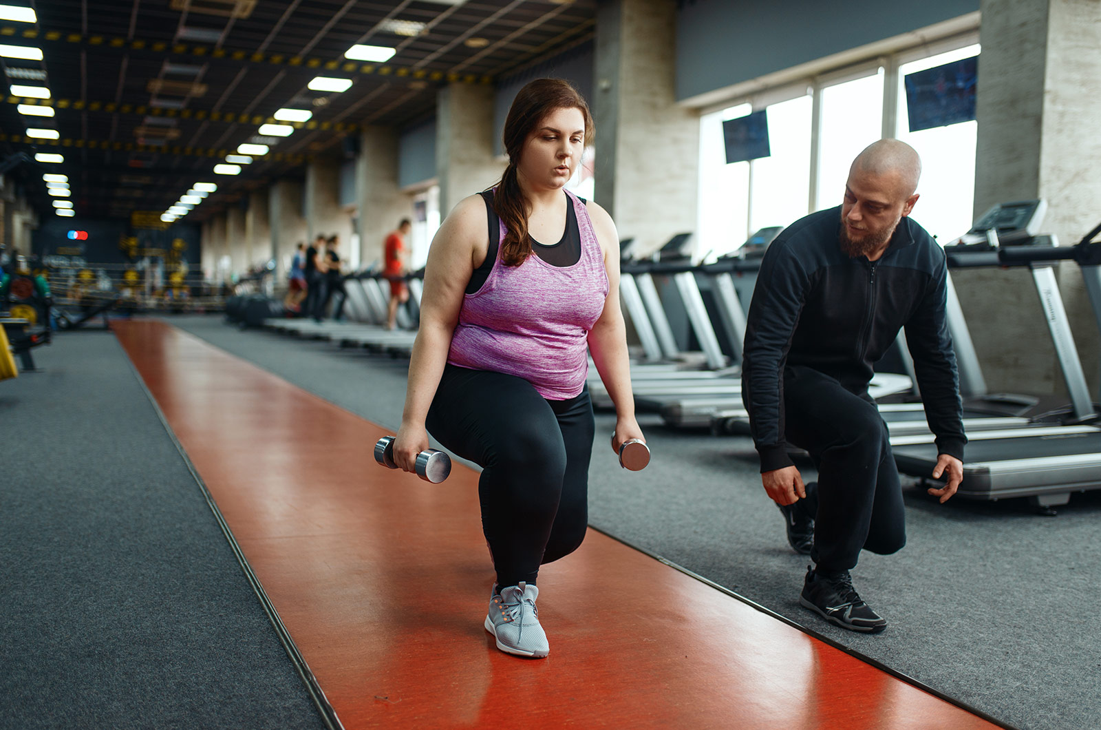Übergewichtige Frau mit Trainer, Frau bei den Kniebeugen mit Hanteln, Training im Fitnessstudio