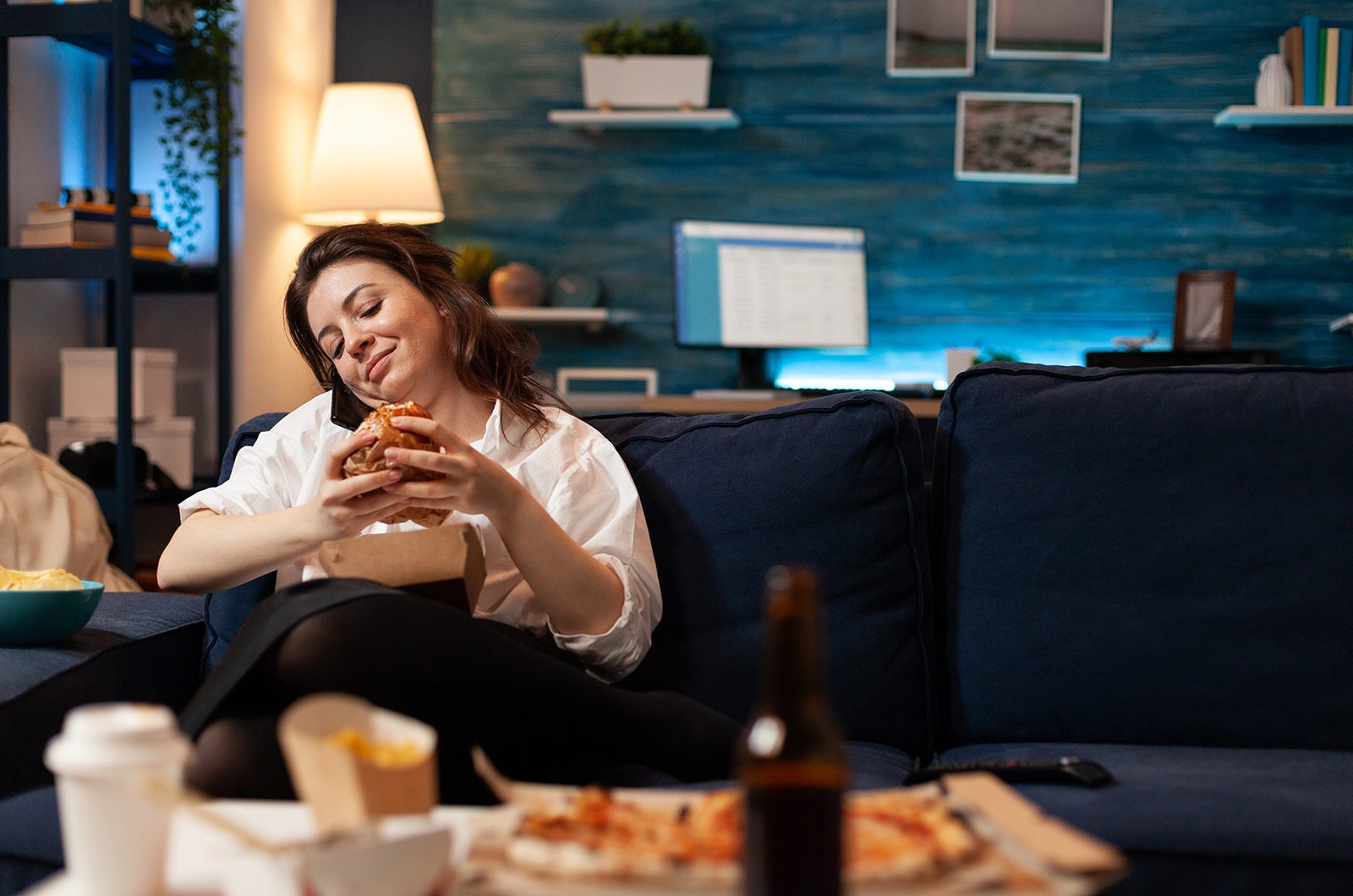 Frau sitzt abends entspannt auf der Couch im Wohnzimmer, telefoniert mit Freunden, während sie einen Burger in den Händen hält, auf dem Tisch davor stehen Pommes, Pizza und eine Flasche Bier