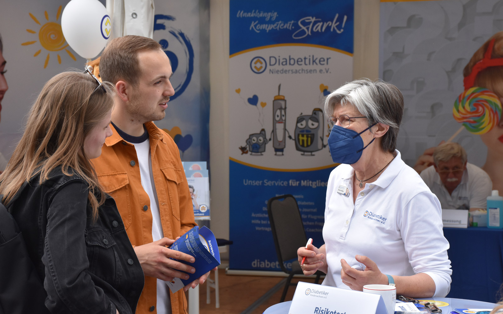 Eine Frau mit Diabetiker Niedersachsen-Polo-Shirt im Gespräch mit einem jungen Paar vor einem Infozelt