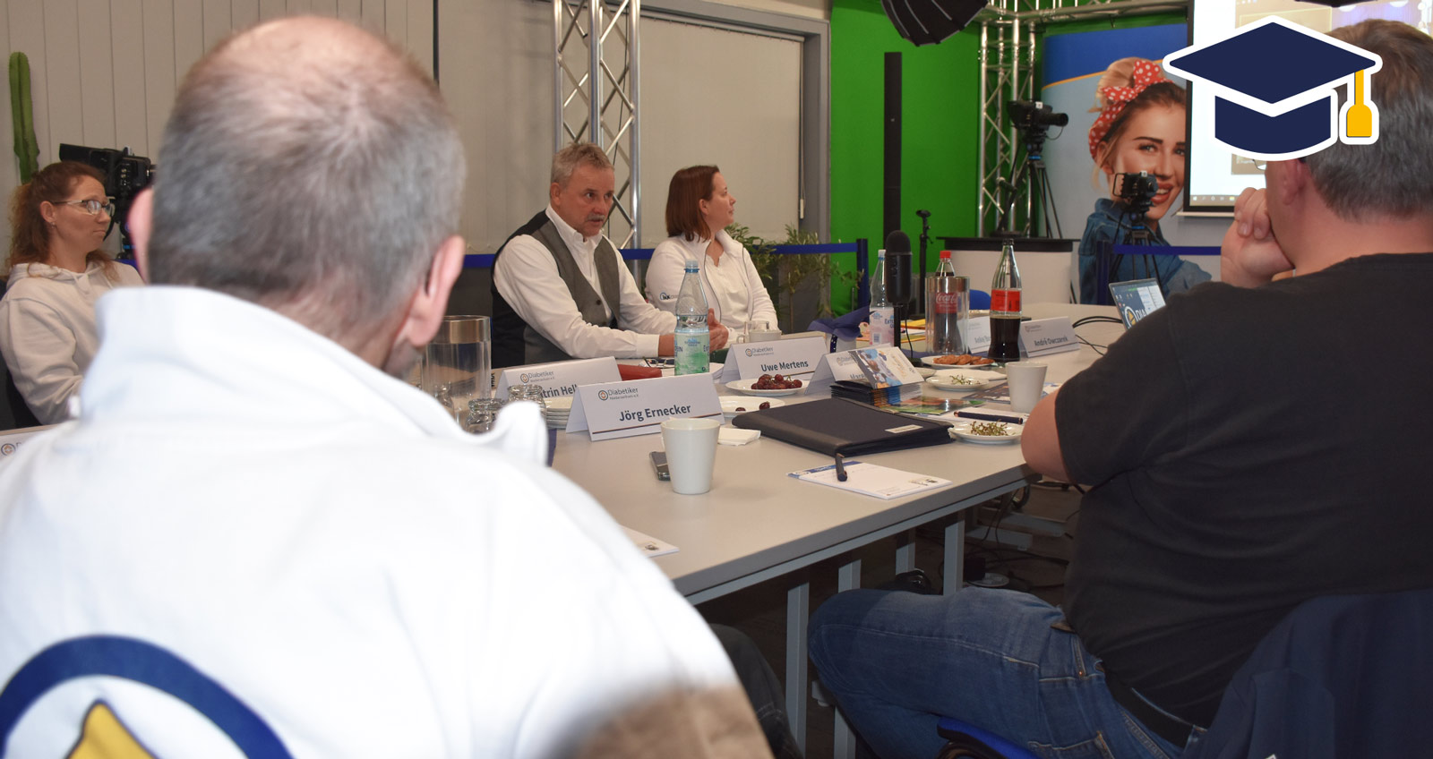 Eine Gruppe von Menschen um einen Tisch, im Hintergund eine Leinwand mit zugeschalteter Videokonferenz. Ein Mann mit Weste referiert. Oben rechts ein blau-gelber Doktorhut im Stil eines Senderlogos