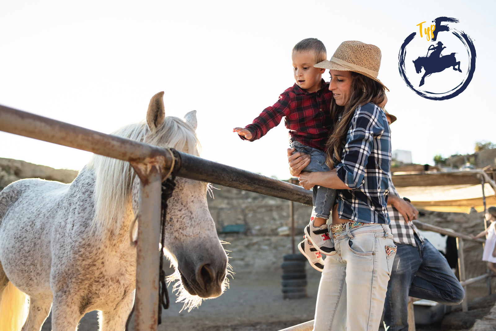Eine Frau hält einen kleinene Jungen auf dem Arm, der über einen Zaun hinweg ein weisses Pferd steichelt