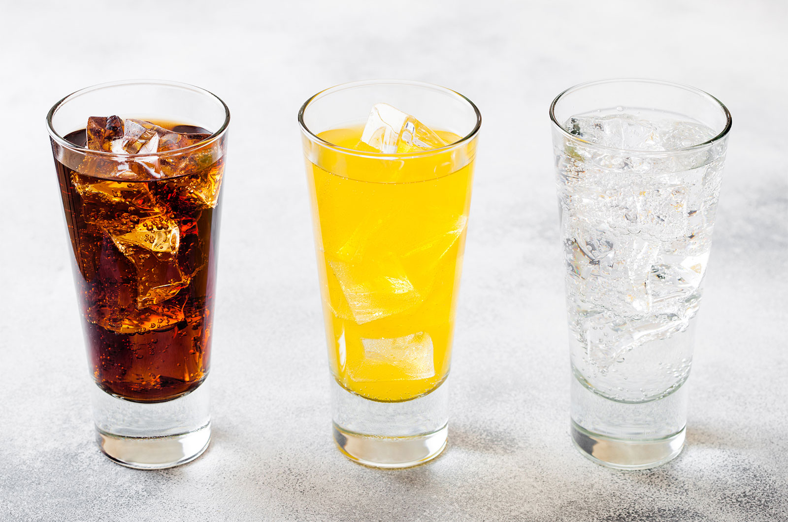 Trinkgläser gefüllt mit Cola, Orangenlimonade und Wasser, mit Eiswürfeln stehend auf einem Stein-Küchentisch