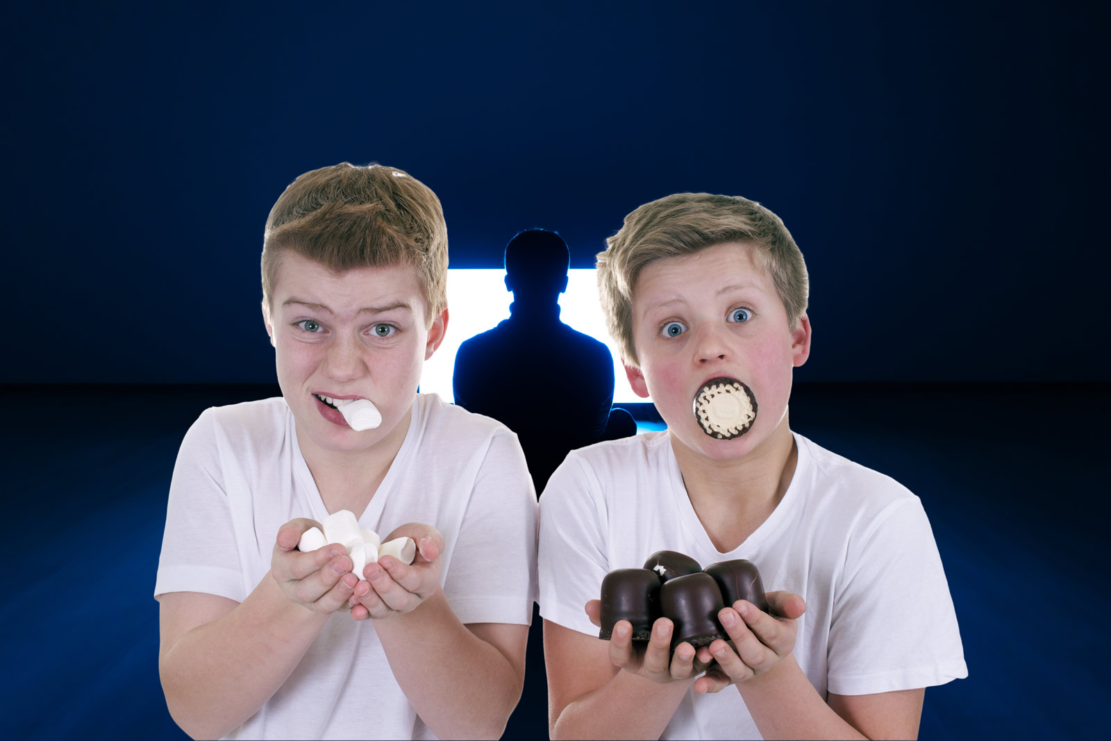 Zwei Jungs stopfen sich mit Süßigkeiten voll. Im Hintergrund läuft ein Fernseher auf den gebannt eine Person starrt.