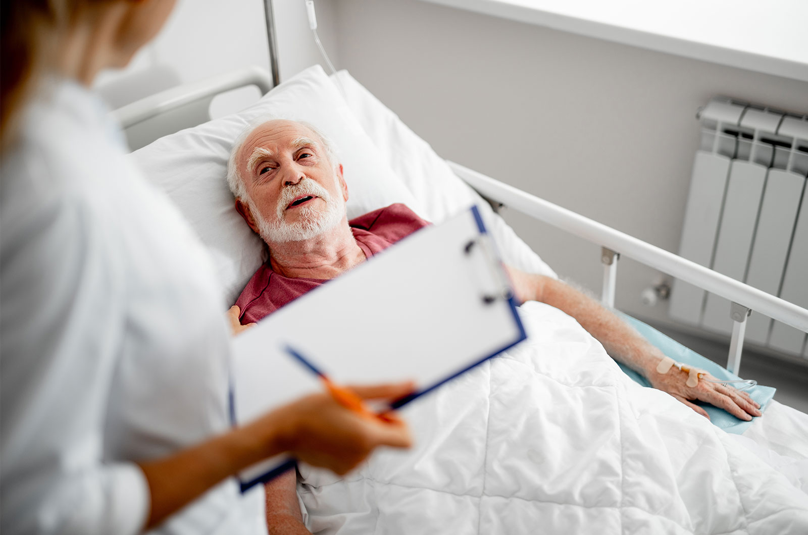 älterer Mann, der im Krankenhauszimmer im Gespräch mit seiner Ärztin über den weiteren Krankenhausaufenthalt spricht 