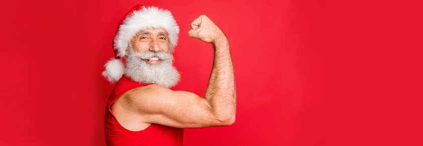 älterer Mann, mit rotem T-Shirt, Weihnachtsmannmütze, weißem Bart, zeigt die trainierten Armmuskeln 