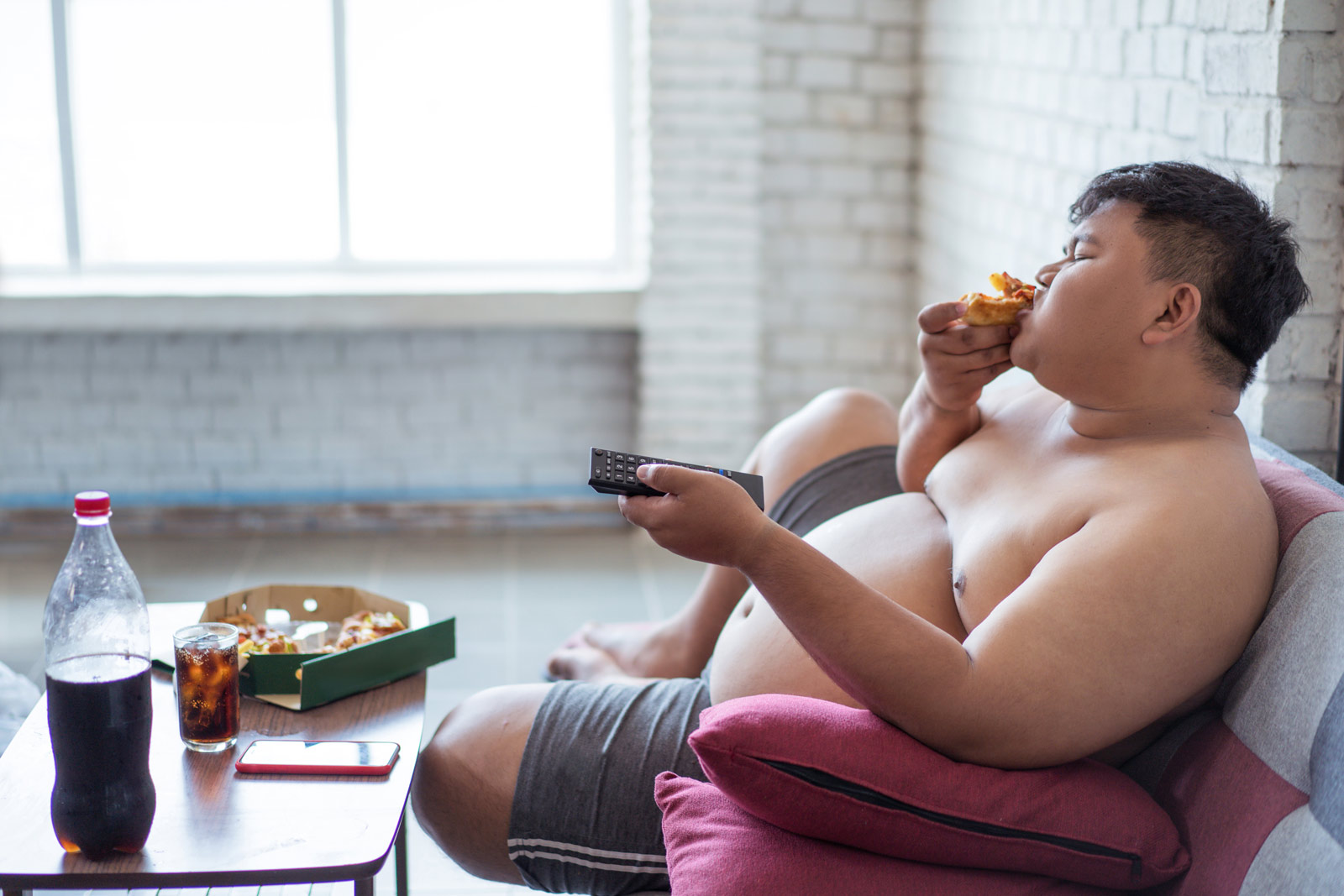 Ein übergewichtiger junger Mann sitzt vor dem Fernseher und isst Pizza