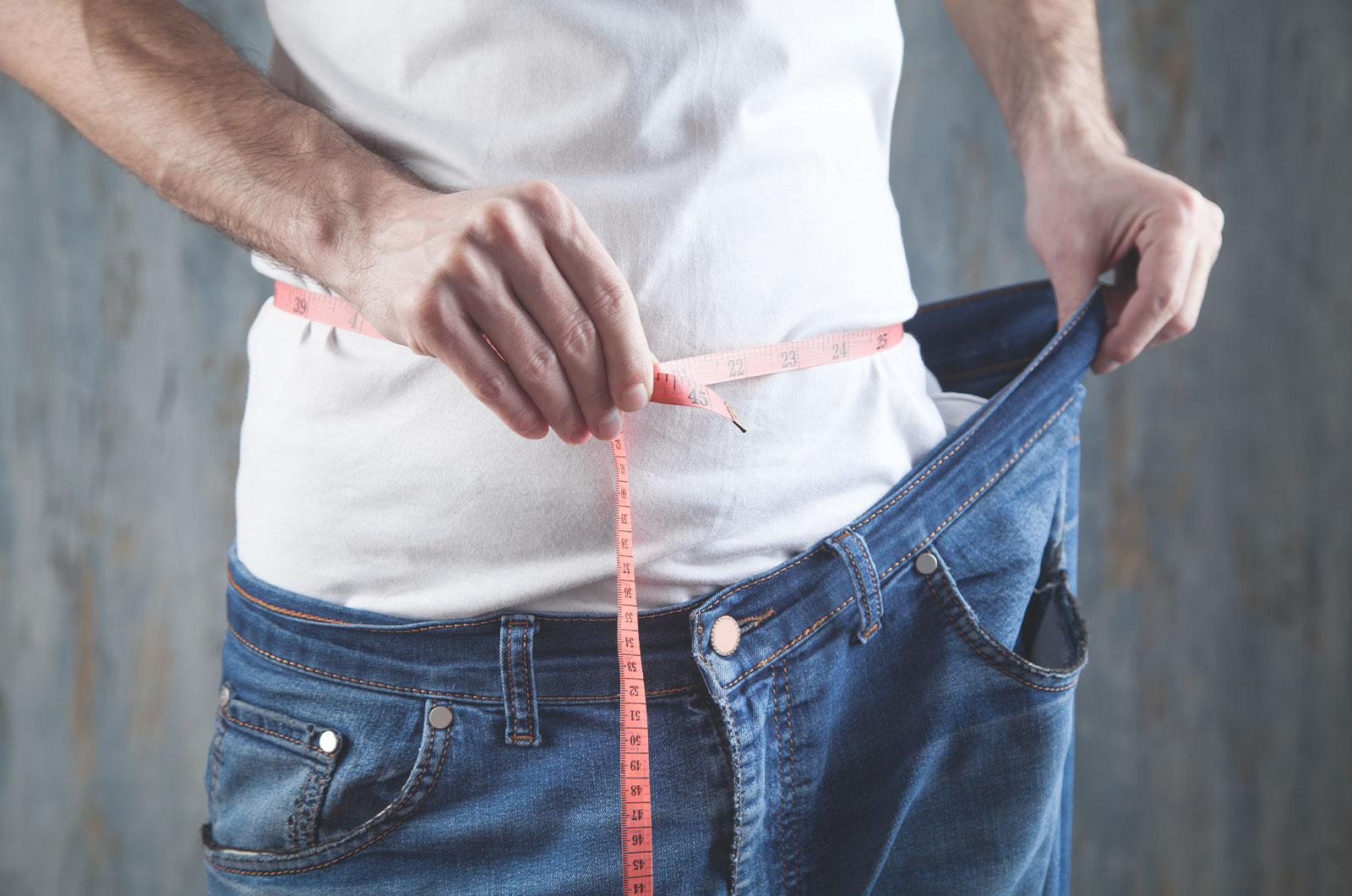 Mann misst seinen Bauchumfang, zeigt die Gewichtsabhame und die viel zu weite Jeans