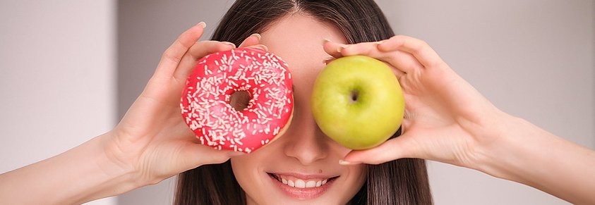 Eine Frau hält sich mit einem Donut und einem Apfel jeweils die Augen zu