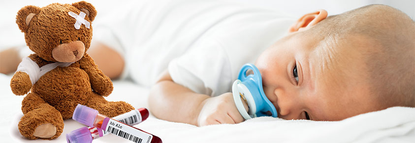 Porträt eines bezaubernd süßen Babys auf dem Bauch in einem weißen Bettchen liegend, im Vordergrund ein Teddybär mit bandagierter Pfote und Pflaster auf dem Kopf und einem Hämoglobin A1c Bluttest